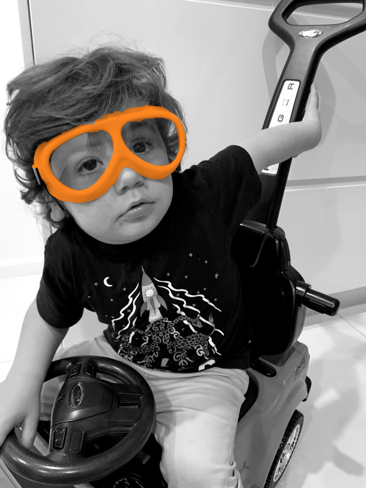 Toddler in orange goggles on a ride on car, black and white with orange goggles