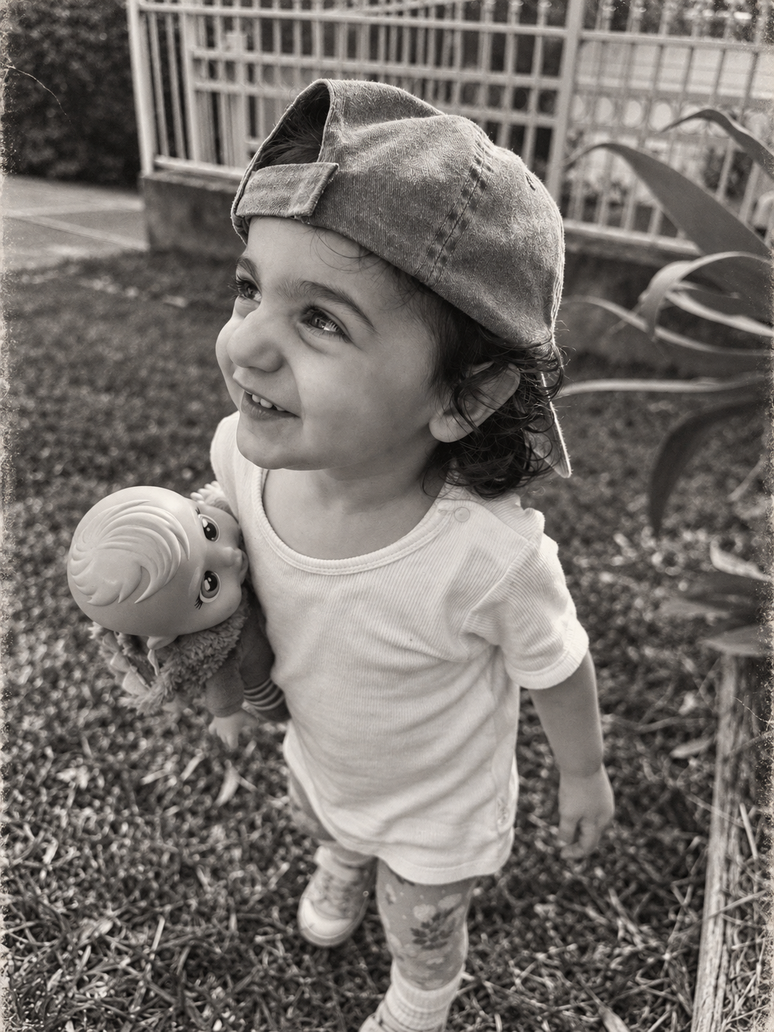 Toddler in a backwards cap holding a doll in the garden, black and white